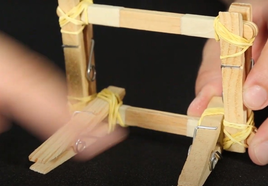 Usando um elástico em cada prendedor de roupas, que formou a base de apoio, enrole e prenda, cruzando em formato de “X”, os prendedores e o conjunto de palitos.