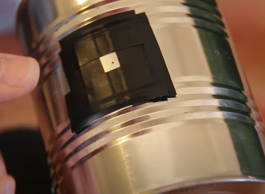 Fixe o pedaço de alumínio furado sobre o furo feito na lata (pelo lado de fora),estes precisam estar alinhados para que o furo menor não esteja obstruído pela parede da lata. Utilize fita isolante para prender e vedar todos os lados da peça de alumínio de forma que a luz só possa entrar pelo furo feito com agulha.